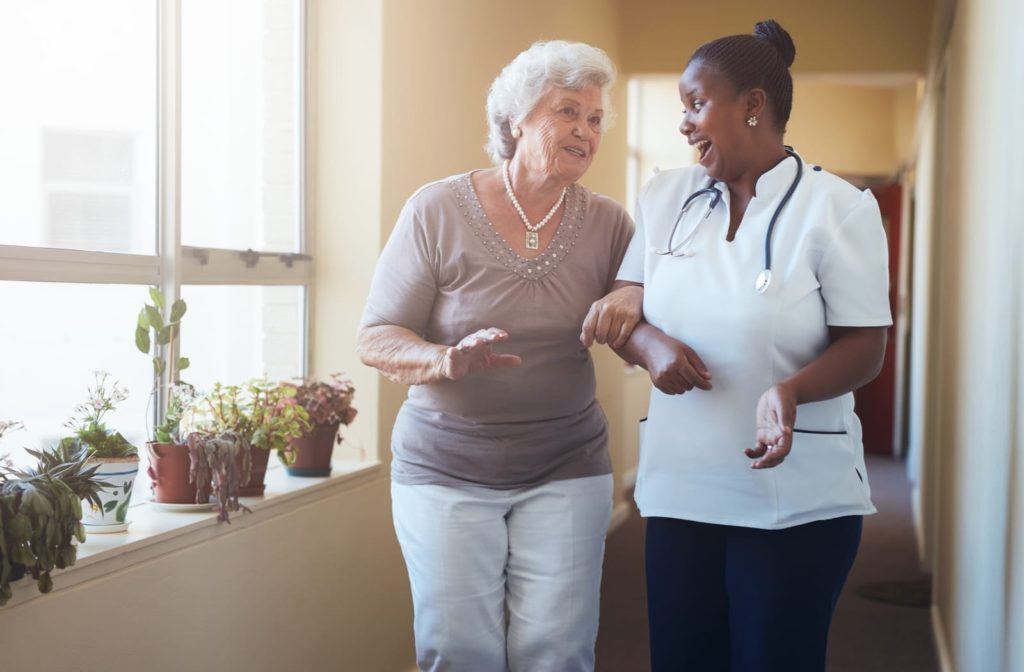 A smiling senior woman receives help walking down a hallway from a friendly health care worker