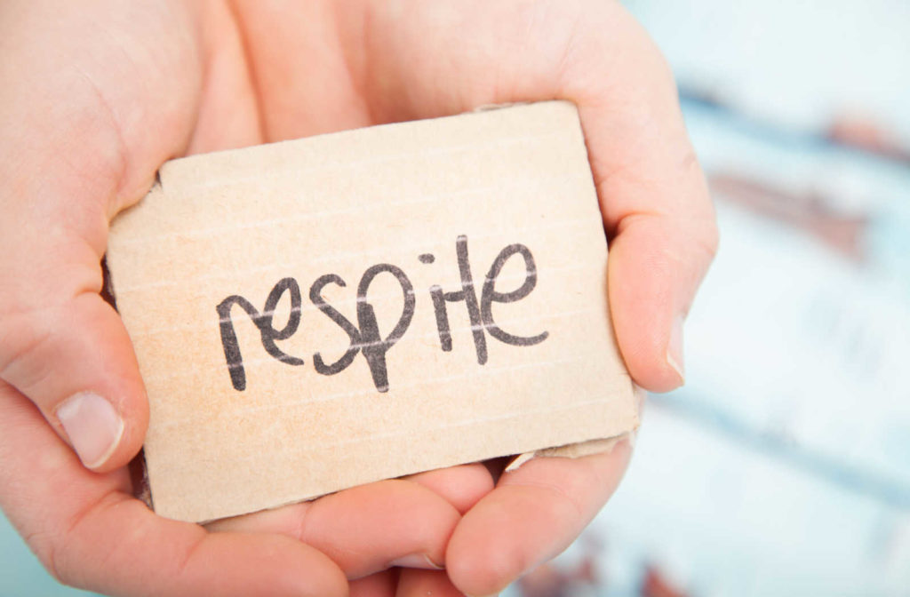 Close up of two hands holding a wooden block with the word respite written on it.