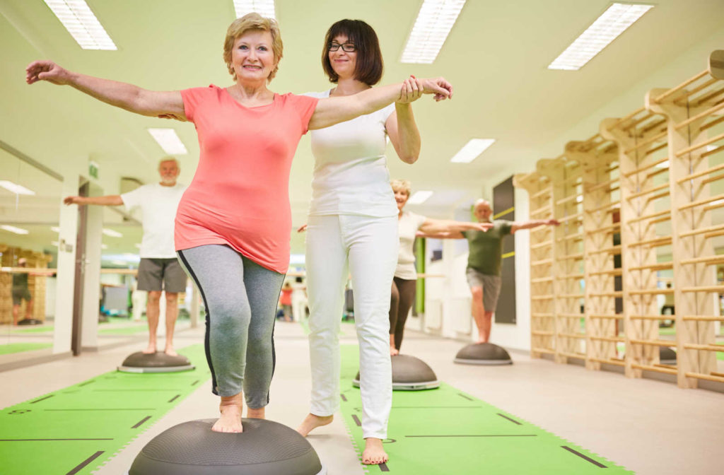 A mature woman balancing on a bosu ball in an exercise class being assisted by the trainer.