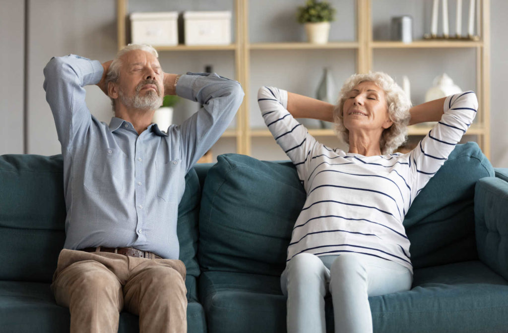 A senior couple sitting on a sofa doing a chest stretch exercise.