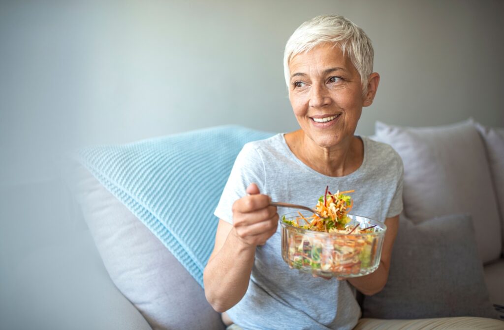 A senior sitting on a couch eats a giant salad full of ingredients that don’t aggravate their dentures