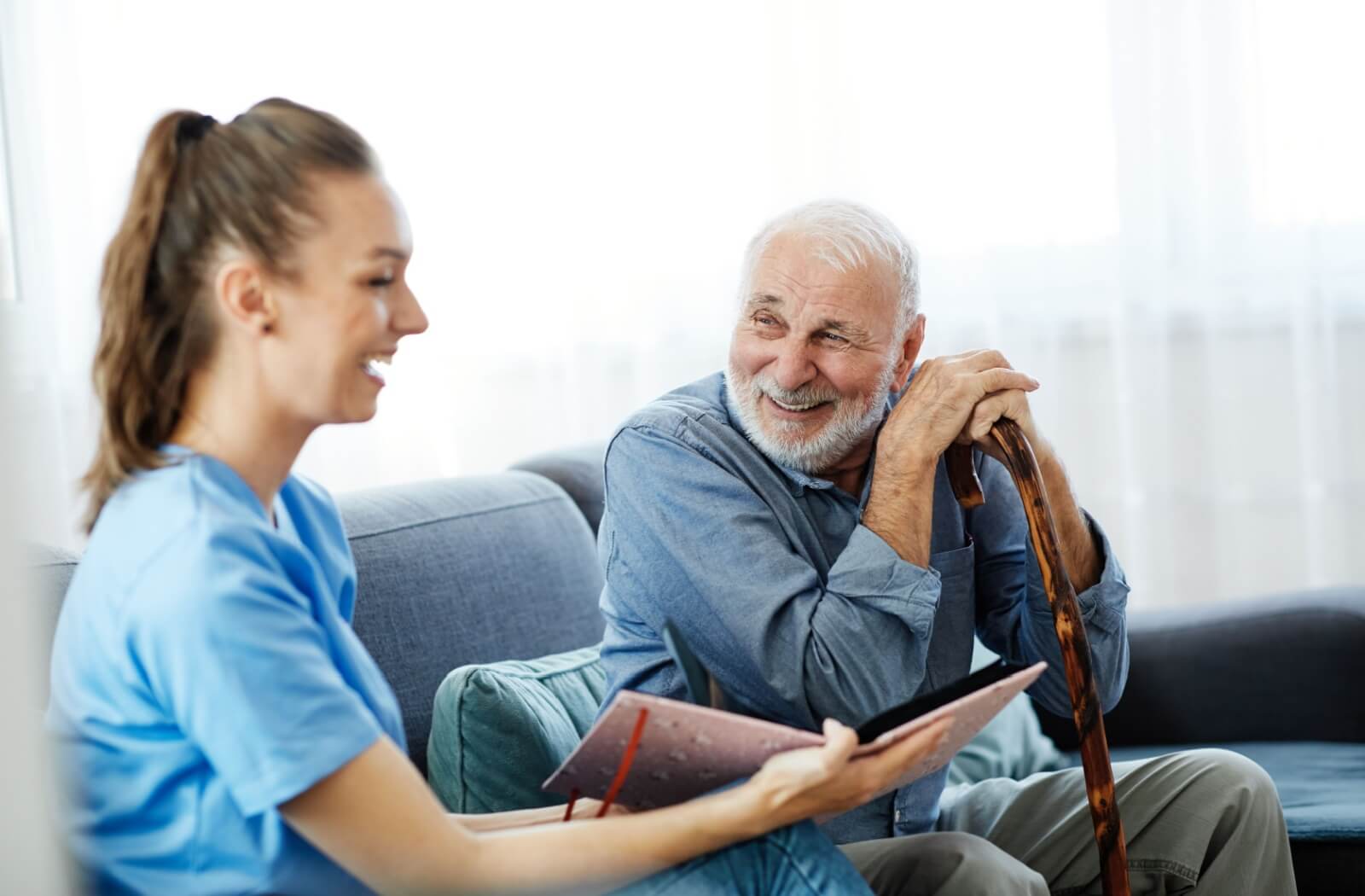 A senior man and a caregiver in memory care sitting on a couch looking at a photo album and smiling.