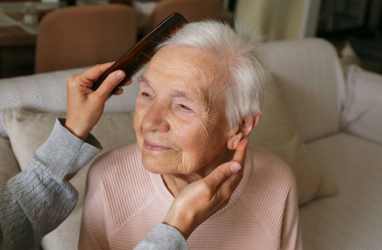 A person gently combs a content senior's soft, white hair.