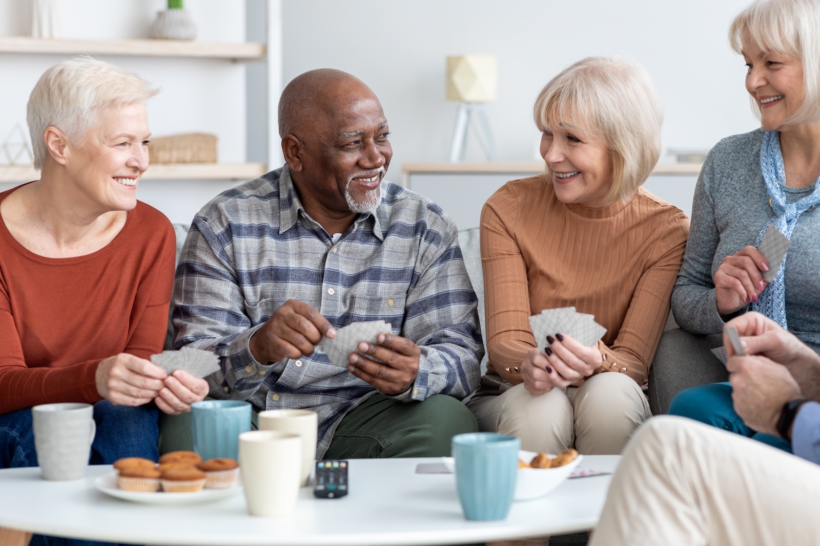 Group of older adults sitting around a table, drinking tea, eating snacks, and playing cards.