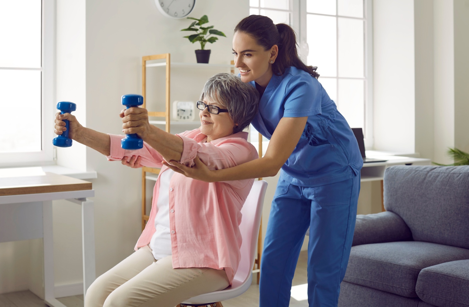 A young nurse in scrubs assists a senior sitting in a chair exercising with lightweight dumbells.