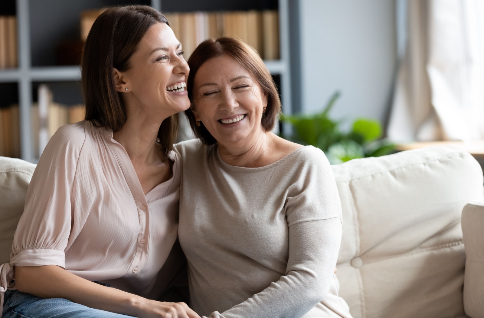 A young adult sitting on a couch hugging and smiling their senior mother.