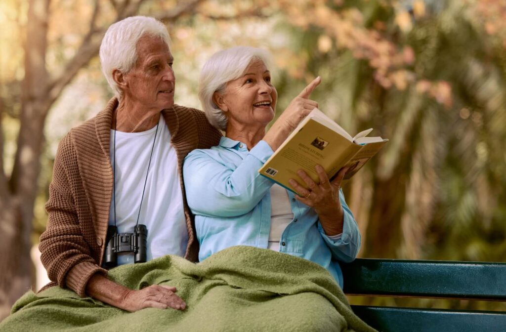 A senior couple bird watches on a bench together, one holding a book and one with binoculars.