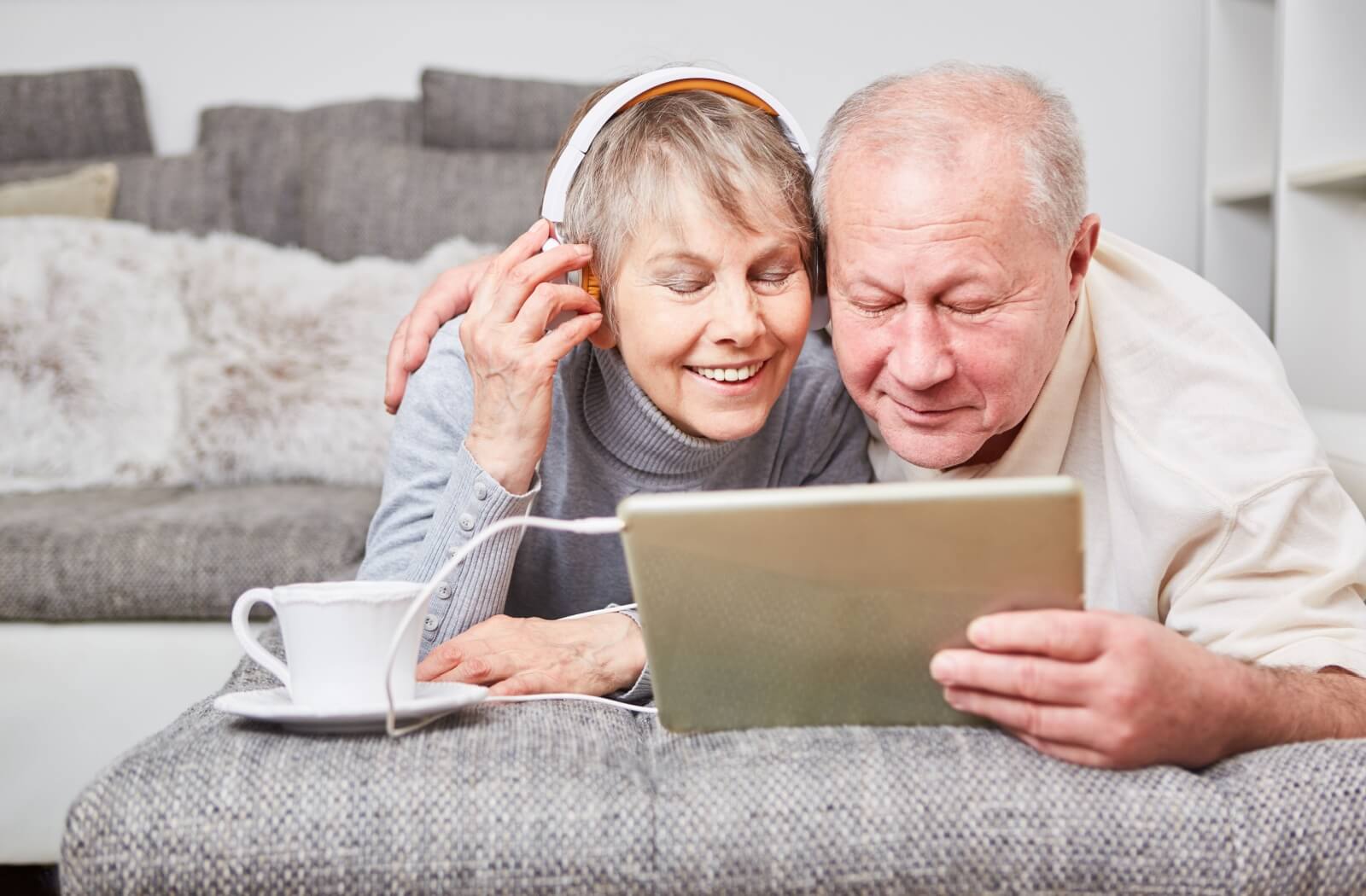 Two seniors relax and listen to an audiobook together with headphones linked to a tablet.