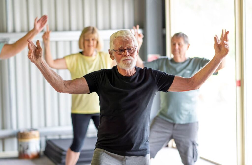 Three seniors participate in a tai chi class provided by their senior living community, holding their arms up in a pose
