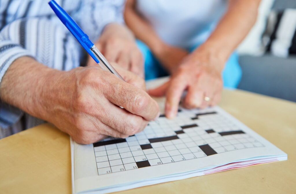 Close-up of senior hands filling in a crossword puzzle with a blue pen while another person points to a clue